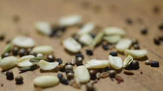 A close-up view of various seeds scattered on a wooden surface, including cumin, coriander, fennel, and black mustard seeds, creating a diverse texture and pattern.