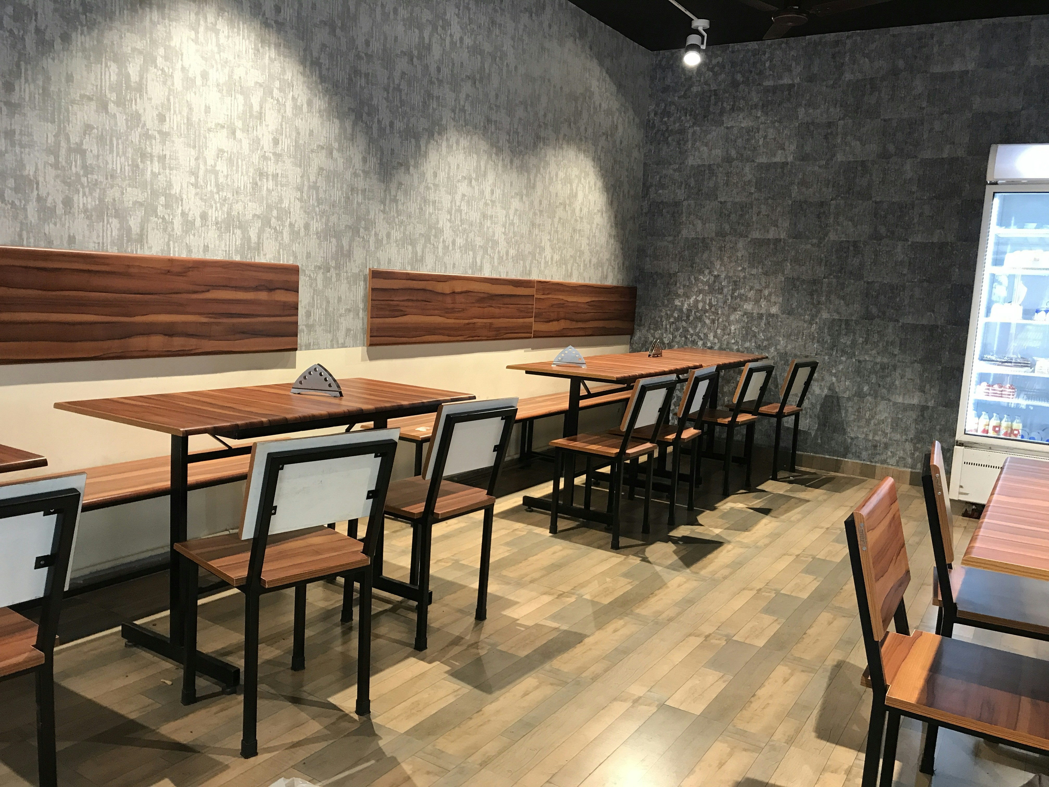 An empty cafe or restaurant area with wooden tables and chairs arranged in rows. The walls have a textured gray finish, and there is a well-lit refrigerator on the right side containing beverages.