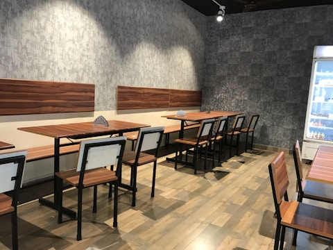 An empty cafe or restaurant area with wooden tables and chairs arranged in rows. The walls have a textured gray finish, and there is a well-lit refrigerator on the right side containing beverages.