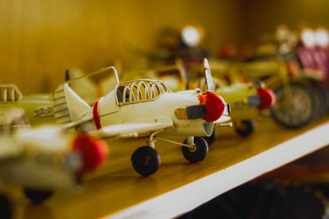 A collection of model airplanes is neatly arranged on a shelf. The focus is on a small vintage-style airplane with a red nose cone and propeller, set against a blurred background of other similar models. The lighting casts a warm, nostalgic glow over the scene.