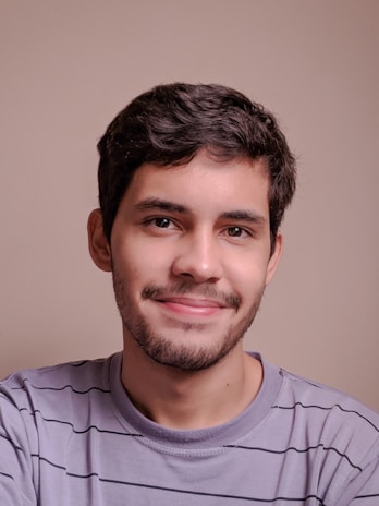 Close-up of a young man smiling gently, wearing a soft purple shirt, with a sandy background.