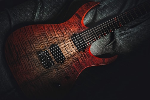 A beautifully crafted electric guitar with a rich mahogany finish and intricate wood grain patterns is seen resting against a dark fabric background. The guitar's body features shiny black pickups and a set of six strings extending across the fretboard.