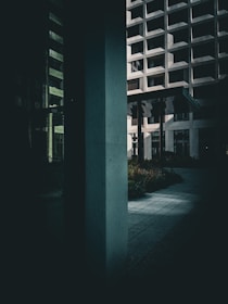 A modern architectural scene with geometric patterns and shadows cast by a multi-story building. The environment features a strong vertical column in the foreground and an open walkway surrounded by landscaped greenery. The image captures a blend of concrete and glass structures, with a play of light and dark areas.