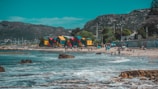 Vibrant, colorful beach huts in green, red, yellow, and blue line the sandy shore beside a picturesque ocean. People enjoy the sunny day, some relaxing with umbrellas while others play in the waves. The background features a mountainous landscape under a clear blue sky, adding a serene natural backdrop.