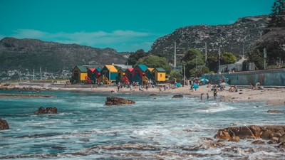 Vibrant, colorful beach huts in green, red, yellow, and blue line the sandy shore beside a picturesque ocean. People enjoy the sunny day, some relaxing with umbrellas while others play in the waves. The background features a mountainous landscape under a clear blue sky, adding a serene natural backdrop.