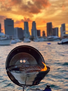 A grill with flames sits by a waterfront, with a city skyline in the background against a dramatic sunset. The vibrant sky features hues of orange and yellow, contrasting with the dark silhouettes of skyscrapers.