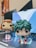 Two stylized figures are positioned on top of a laptop keyboard. The figures, likely based on animated characters, have exaggerated features such as large eyes and unique hairstyles. The background comprises a blurred view of a room with a laptop screen displaying a colorful wallpaper.