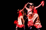 A lively group of dancers captured mid-performance. The dancers wear vibrant traditional dresses in red and gold, with dynamic movements. Their arms are raised, holding colorful fabric, against a stark black background.