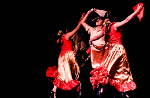 A lively group of dancers captured mid-performance. The dancers wear vibrant traditional dresses in red and gold, with dynamic movements. Their arms are raised, holding colorful fabric, against a stark black background.