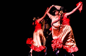 A lively group of dancers captured mid-performance. The dancers wear vibrant traditional dresses in red and gold, with dynamic movements. Their arms are raised, holding colorful fabric, against a stark black background.