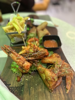 A wooden board with various types of fried food including prawns, chicken wings, French fries, and dipping sauce in a small black dish. The board is garnished with fresh green lettuce leaves. There is a metallic basket holding the fries. The background is slightly blurred with hints of people and tables.