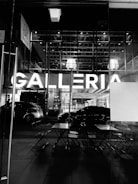A black and white photo features the entrance of a modern shopping center. The word 'GALLERIA' is prominently illuminated in large letters. In the foreground, there are folded chairs and tables, suggesting an outdoor seating area. Cars are parked nearby, reflecting the urban setting. The glass facade reveals a glimpse of the interior lights and ceiling structure.