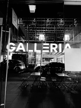 A black and white photo features the entrance of a modern shopping center. The word 'GALLERIA' is prominently illuminated in large letters. In the foreground, there are folded chairs and tables, suggesting an outdoor seating area. Cars are parked nearby, reflecting the urban setting. The glass facade reveals a glimpse of the interior lights and ceiling structure.