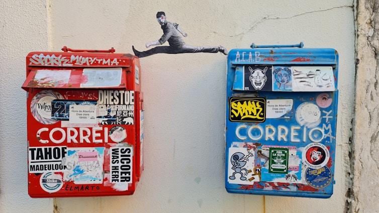 Two colorful mailboxes, one red and the other blue, are mounted on a wall. Both are covered in various stickers and graffiti. Above the mailboxes is a piece of street art depicting a leaping person with a black hat and blue face paint.