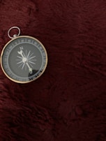 A silver compass with a circular face rests on a deep red, textured surface. The compass needle points towards the north, with cardinal directions clearly marked around the edge.