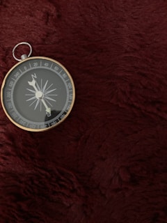 A silver compass with a circular face rests on a deep red, textured surface. The compass needle points towards the north, with cardinal directions clearly marked around the edge.