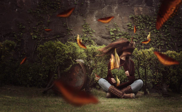 A person sits cross-legged on the grass holding a mirror frame with flames emerging from it. Their long hair flows dramatically, blending into the fiery elements surrounding them. Orange and red butterfly-like shapes hover in the air. The background features a stone wall covered in ivy and small bushes.