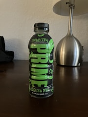 A black and green bottle of Prime Hydration Drink is placed on a dark wooden surface. The label reads 'Glowberry' and '16.9 FL OZ (500 mL)'. In the background, a metallic lamp with a white shade is visible.