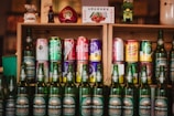 A collection of various drink bottles and cans is displayed on wooden shelves. The shelves contain a variety of soda cans with diverse branding, and in front of them are several green bottles labeled Taiwan Beer. The setup is decorated with small figurines and a decorative item shaped like a heart.