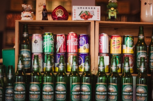 A collection of various drink bottles and cans is displayed on wooden shelves. The shelves contain a variety of soda cans with diverse branding, and in front of them are several green bottles labeled Taiwan Beer. The setup is decorated with small figurines and a decorative item shaped like a heart.