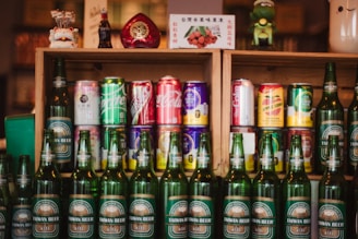 A collection of various drink bottles and cans is displayed on wooden shelves. The shelves contain a variety of soda cans with diverse branding, and in front of them are several green bottles labeled Taiwan Beer. The setup is decorated with small figurines and a decorative item shaped like a heart.
