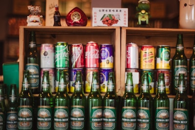 A collection of various drink bottles and cans is displayed on wooden shelves. The shelves contain a variety of soda cans with diverse branding, and in front of them are several green bottles labeled Taiwan Beer. The setup is decorated with small figurines and a decorative item shaped like a heart.