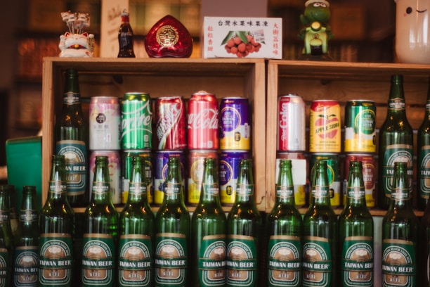 A collection of various drink bottles and cans is displayed on wooden shelves. The shelves contain a variety of soda cans with diverse branding, and in front of them are several green bottles labeled Taiwan Beer. The setup is decorated with small figurines and a decorative item shaped like a heart.
