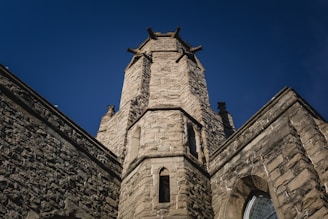 A stone tower rises against a clear blue sky. The structure features intricate masonry with narrow, arched windows and decorative elements at the top. The tower is part of a larger stone building that includes additional architectural details such as angled buttresses and leaded glass windows.