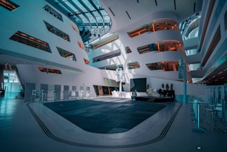 A modern, spacious interior with futuristic architecture featuring curved walls and large windows. The space has a high ceiling with ample natural light coming through. There are a few people sitting in the center on sleek furniture, surrounded by minimalistic, contemporary decor, including a series of elevated walkways and alcoves with reflective surfaces.