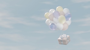A cluster of pastel-colored balloons is carrying a gift box with a ribbon through a light blue sky with soft clouds.