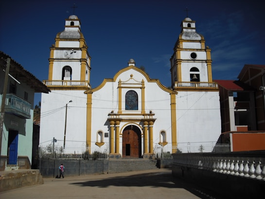 A large, white church with two bell towers and yellow trim stands prominently against a clear blue sky. The church features a large wooden door and a stained-glass window above it. In front of the church, there is a person walking along a paved path. The architecture shows colonial influences with classical decorative elements.