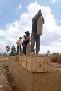 Three individuals are engaged in a manual construction task, standing on a wooden frame filled with earth. They are holding large wooden implements, appearing to compact the material. The setting is outdoors with a partly cloudy sky and some greenery in the background.