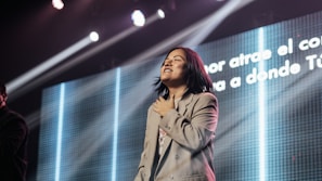 A person appears to be on stage with a hand over the heart, possibly in a moment of emotional expression. Bright lights shine down, creating an atmospheric effect. A large screen in the background displays text in Spanish.