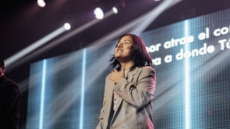 A person appears to be on stage with a hand over the heart, possibly in a moment of emotional expression. Bright lights shine down, creating an atmospheric effect. A large screen in the background displays text in Spanish.