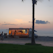 A food truck is parked on a pebbly beach during sunset. The sky is painted with soft hues of pink and orange, and a couple of clouds dot the horizon. A palm tree is silhouetted in the foreground, and a group of people stand near the food truck, illuminated by its warm lights.