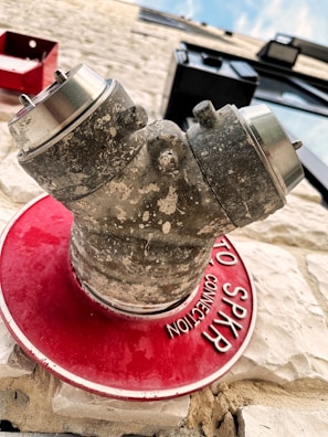 A weathered and textured metallic pipe fixture is mounted on a white textured brick wall. The fixture appears to be a fire department connection, as indicated by the red circular plate with white text that partially reads 'SPKR CONNECTION'. The background includes a glimpse of the sky and part of the building structure, with a black window frame in view.