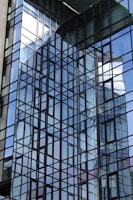 A modern glass building facade with a grid-like pattern of reflective windows. The structure showcases a complex interplay of light and reflections, with surrounding architecture mirrored in the glass. The angular lines create a dynamic visual effect.