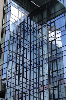A modern glass building facade with a grid-like pattern of reflective windows. The structure showcases a complex interplay of light and reflections, with surrounding architecture mirrored in the glass. The angular lines create a dynamic visual effect.
