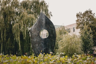A large, abstract, dark metal sculpture with an oval shape and a central circular hole is situated outdoors in a lush green environment. Surrounding the sculpture are trees with dense foliage and possibly some buildings in the background. The scene also includes a hedge or shrubs in the foreground.