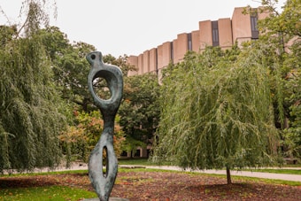 A modern abstract sculpture is situated in a landscaped area with lush green trees. The sculpture features organic, flowing shapes with hollows in its form, made of a weathered green material. Behind the sculpture, a large, uniquely designed building with an angular facade is partially visible. The surroundings include a path and well-maintained garden spaces.