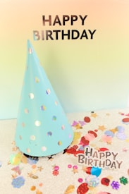 A light blue party hat with iridescent polka dots is placed on a silver glittery surface that is scattered with colorful confetti and shiny cutouts. The words 'Happy Birthday' are prominently displayed in bold black letters at the top of the image against a gradient background.