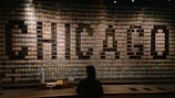 A large wall composed of numerous colorful items arranged meticulously to spell out the word 'CHICAGO'. Below the wall, there is a counter with cups, napkins, and other items visible. The lighting focuses on the central part of the display, creating a dramatic effect. A person is standing in front of the counter, facing the wall.