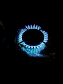 A gas stove burner with a blue flame emitting from its circular openings. The flame is vibrant, with visible hints of orange around the edges, and the surrounding area is dark, highlighting the glow of the fire.