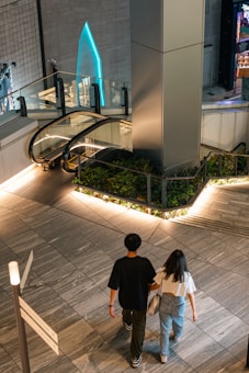 A modern interior space features a sleek escalator surrounded by glass railings and greenery. Two people walk in the foreground on a polished tile floor, heading towards the escalator. Ambient lighting creates a warm and inviting atmosphere, with a futuristic architectural design highlighted in the background.