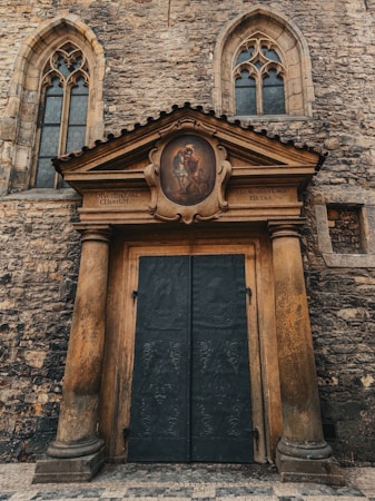 A historic stone building with two arched windows featuring intricate stained glass details. Below the windows, there is an ornate wooden door framed by two large, textured columns. Above the door is a pediment with a circular painting depicting a religious scene. Latin inscriptions are visible on the facade.