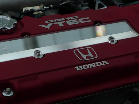 A close-up view of a car engine cover featuring the red and silver branding of a Honda DOHC VTEC engine. The cover has a metallic Honda logo and several screws visible.