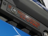 A close-up view of an engine cover displaying the logo of a luxury car brand and the text '3.2 VTEC'. The cover is primarily black with red accents and is part of a shiny blue surface below it.