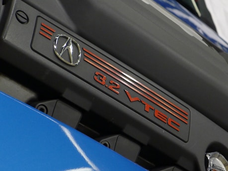 A close-up view of an engine cover displaying the logo of a luxury car brand and the text '3.2 VTEC'. The cover is primarily black with red accents and is part of a shiny blue surface below it.