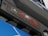 A close-up view of an engine cover displaying the logo of a luxury car brand and the text '3.2 VTEC'. The cover is primarily black with red accents and is part of a shiny blue surface below it.