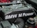 A detailed view of a BMW engine component featuring carbon fiber material with the text 'BMW M Power' prominently displayed. Various engine parts and components are visible around the main object, including cables, hoses, and connectors.